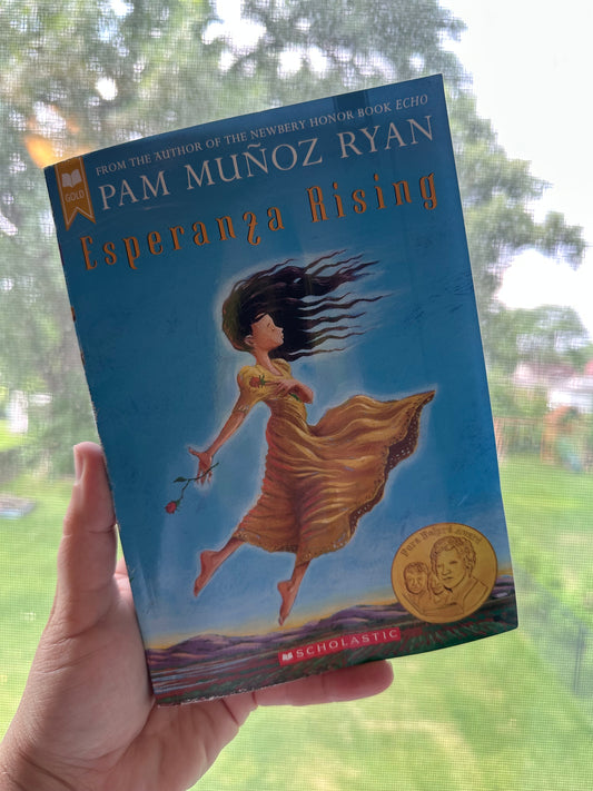 Book titled 'Esperanza Rising' held in front of a window with trees in the background